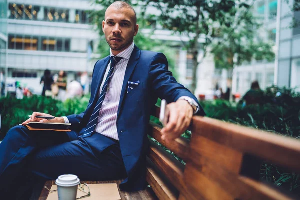 Portrait of Caucasian adult student in formal clothing sitting at urban setting and spending time for planning university project,intelligent professor 30s holding education notepad looking at camera