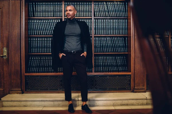 モダンなオフィスの快適なオフィスで茶色の木製の本棚に対してポケットに手で立って離れて見て 考えてスタイリッシュなエレガントな服の高級感のある執行者の男の低角度 — ストック写真