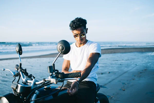 身穿太阳镜的时尚男子坐在停在空旷沙滩边的摩托车旁 蓝天上的海滨波涛汹涌 — 图库照片