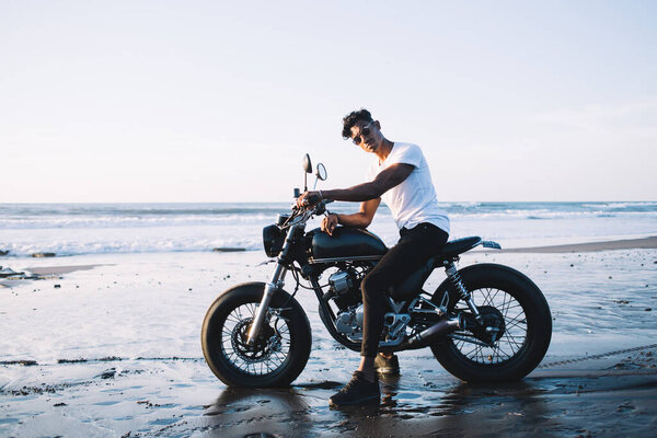 Secluded ethnic young confident keen man in sunglasses resting on modern motorbike and enjoying freedom being out on sea beach around sunset