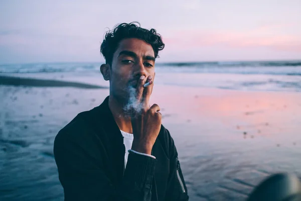 暗いジャケットを身に着けているタバコを保持し 煙を吐き出すと日没の夜に海辺のカメラを見てフラストレーションヒスパニック男性 — ストック写真