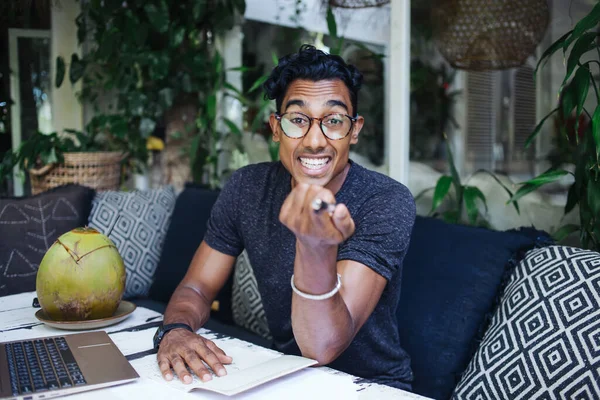 Excited youthful Hispanic college student in glasses pointing and looking at camera while preparing homework and sitting at table in terrace