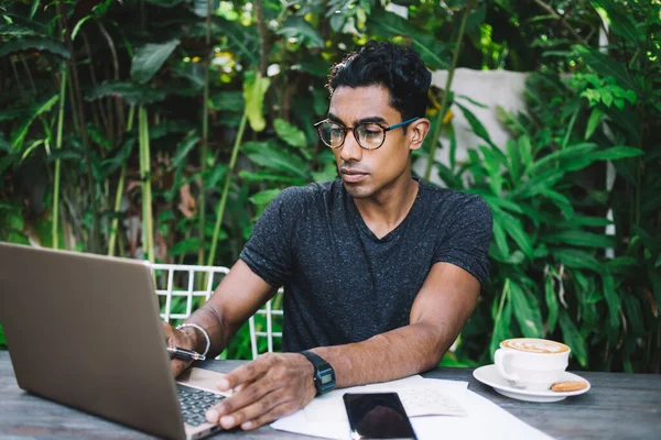 Diligent ethnic man in eyeglasses and casual outfit using laptop while sitting at table with documents and coffee cup at street cafe
