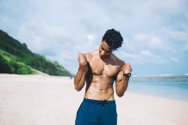 黒髪若いです民族の自信のある男性で腕時計で青のショートパンツは筋肉を示すジェスチャーで広々としたビーチに立って — ストック写真