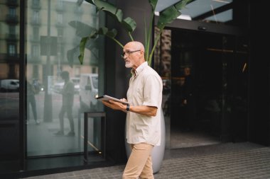 Düşünceli, günlük giysiler ve gözlüklü bir adamın şehir caddesinde yürümesi ve elinde tabletle dolaşması.