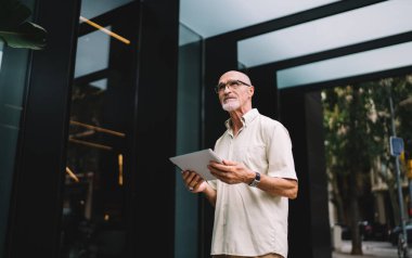 Gündelik giysiler ve gözlüklerle şehir caddesindeki modern binanın yanındaki kaldırımda duran ve tablet kullanan dalgın erkek.