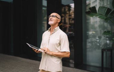 Düşünceli son sınıf öğrencisi, günlük kıyafetleri ve gözlükleriyle şehir caddesinde duruyor ve tableti elinde tutuyor.