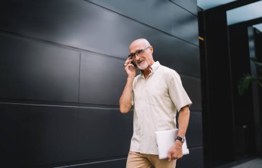 Şehir caddesinde yürüyen ve elinde tabletle akıllı telefondan konuşan sıradan giysili ve gözlüklü mutlu sakallı adamdan.