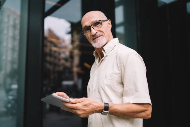 Beyaz tişörtlü ve gözlüklü yaşlı bir adamın şehir caddesinde durup tableti elinde tutması.