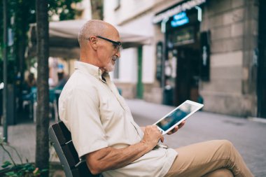 Pozitif sakallı, günlük giysiler ve gözlüklerle caddede bankta dinlenirken tablet kullanan yaşlı erkek yöneticinin yan görüntüsü