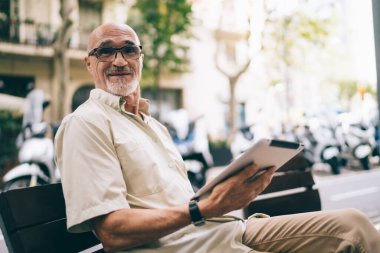 Mutlu, gri sakallı, günlük giysiler içinde, gözlüklü, gülümseyen ve kameraya bakan bir adam.
