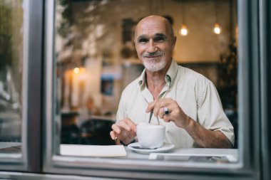 Rahat kafeteryada pencereye yaslanıp kameraya bakarken günlük giysiler içinde gülümseyen ve cappuccino içen yaşlı bir erkek.