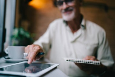 Çağdaş çalışma alanında çalışırken tablet kullanarak ve not defterine internetten bilgi yazarak günlük giysiler içinde gülen kıdemli sakallı yönetici.