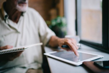 Kafedeki masada çalışırken not defteri ile tablet ekranına dokunan yüzsüz, yaşlı, sakallı iş adamı.