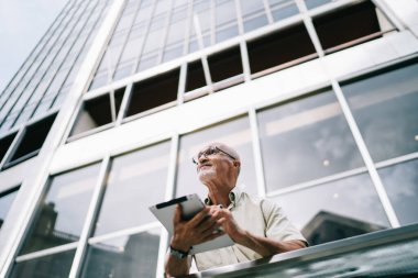 Gözlüklü, gözlüklü, tablet kullanan ve gündüz gözlüklü, modern cam gökdelenin önünde dikilirken yüzünü çeviren yaşlı erkek işçilerden.