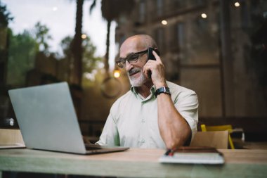 Gözlüklü, cep telefonuyla konuşan ve kafede dizüstü bilgisayarla otururken bilgisayar monitörüne bakan yaşlı, pozitif sakallı bir adam.