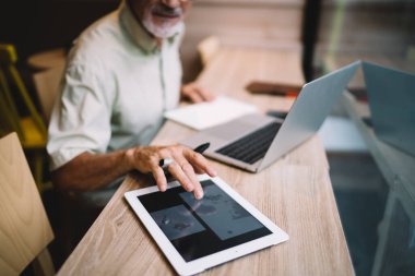 Resmi gömlek içinde tanınmayan sakallı bir erkek. Tabletini karıştırıyor ve yaratıcı çalışma alanında masa başında oturup dizüstü bilgisayarla çalışırken not alıyor.