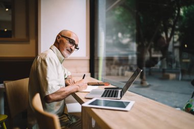Resmi giyim ve gözlük takan, not yazan ve bilgisayarla çalışan, yaratıcı çalışma alanında masa başında tablete bakan konsantre yaşlı girişimcilerin yan görüntüsü