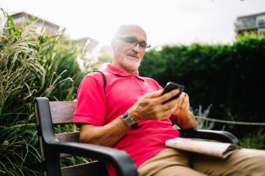 Günlük kıyafetleri ve gözlükleriyle yaşlı sakallı adam yeşil bitkilerin yanında bankta oturuyor ve hafta sonu dinlenirken akıllı telefondan mesajlaşıyor.
