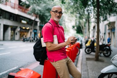 Sakallı, kel, stil sahibi, gözlüklü, sırt çantalı, kaşlarını kaldırmış, akıllı telefon kullanan ve gündüz vakti bisiklet standına yaslanan son sınıf öğrencisinin yan görüntüsü.