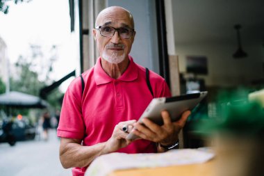 Parlak pembe tişörtlü ve gözlüklü şaşırmış bir erkek. Kaşlarını kaldırmış, tablet üzerinde internete bakıyor. Sokak kafeteryasında oturmuş kameraya bakıyor.