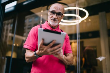 Gündelik giysiler ve gözlüklü, odaklanmış, sakallı bir adamdan tablet üzerinde modern mağazanın cam vitrinine karşı mesaj okuyordu.