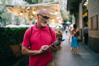Odaklanmış, pembe polo tişörtlü sakallı adam şehir kaldırımında yürürken harita uygulamasında tablet üzerinde arama yapıyor.