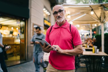 Kafası karışmış, günlük giysiler ve gözlüklü yaşlı bir adam kameraya bakıyor ve tablet üzerinde açık kafeye karşı yön arıyor.