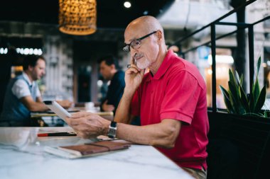 Günlük kıyafet ve gözlüklü yaşlı adam bilgi aramak için tablet kullanıyor ve rahat bir kafede masada oturuyor.