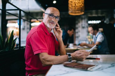 Kel kafalı, sıradan giyinmiş, bilgi aramak için tablet kullanan, kafede oturan ve kameraya bakan pozitif sakallı adamın yan görüntüsü.