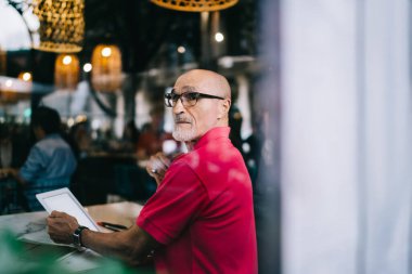 Gözlüklü, sıradan giyinmiş, tablet seyreden, rahat bir kafede oturup gözlerini kaçıran ciddi bir adamın yan görüntüsü.