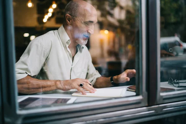 穿着休闲装阅读文章并在组织者中做笔记而独自在创造性工作空间中工作的积极老年留着胡子的男性记者的粗糙玻璃窗侧视图 — 图库照片