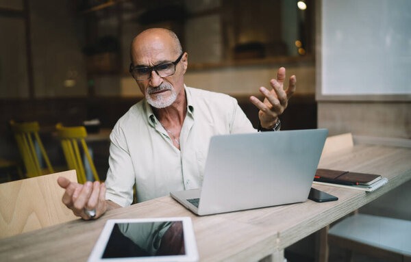 Perplexed frustrated senior entrepreneur in eyeglasses and formal wear working on laptop and looking at tablet while sitting at table in cafe