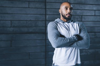 Yakışıklı, kaslı, Afro-Amerikan kel, sakallı, spor kıyafetli, kollarını çapraz yapmış, gri arka plana bakan yetişkin erkek.