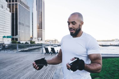 Yakışıklı, kel, spor kıyafetli Afrikalı Amerikalı. Nehir ve şehir merkezinin yakınındaki ahşap iskelede akıllı telefon tutarken kulaklıklarını çıkarıyor ve çözüyordu.