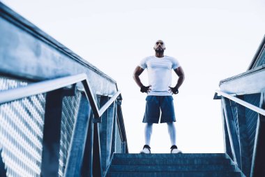Aşağıdan kendine güvenen ve yakışıklı Afro-Amerikan spor giyim koçu ve merdivenlerin başında durup kameraya bakan eldivenler.