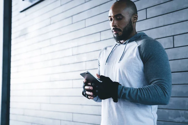 stock image Bearded sporty black male in gloves browsing smartphone while standing alone against gray brick wall in modern fitness gym in daylight