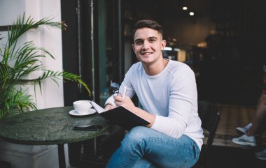 Tatmin olmuş, koyu renk saçlı, gayri resmi kıyafetli, kafede oturmuş kameraya bakarken not defteriyle gülümseyen bir erkek.