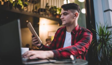 Düşük açılı, gündelik giyinmiş genç bir erkek. Panoda notlar okuyor ve kafede otururken dizüstü bilgisayarda yazıyor.
