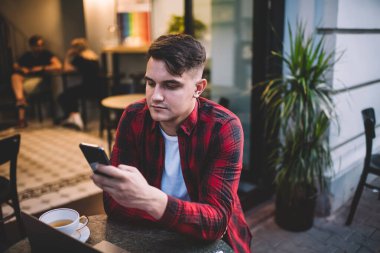 Bir fincan çay içerken günlük kıyafetlerini giymiş, sokak kafeteryasında oturmuş ve cep telefonunu karıştırıyor.