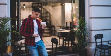 Akıllı telefonlu ve çay bardağıyla günlük giysiler içinde neşeli bir erkek hippi sokak kafesinde ekrana bakıyor.