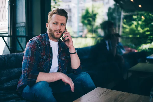 Calm thoughtful bearded male in casual clothes making phone call while sitting on sofa at wooden table on balcony against blurred dark glass wall with reflection