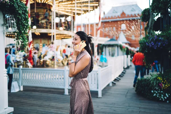 Cheerful millennial woman spending leisure day in amusement park using cellphone gadget for communicate with best friend,happy hipster girl sharing impressions about travel journey during roaming call