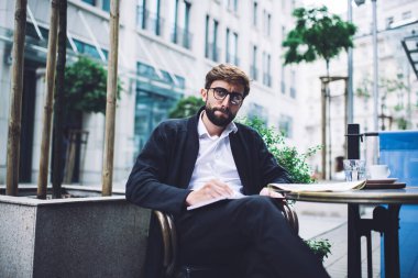 Resmi kıyafetli ve gözlüklü endişeli bir girişimci, kafede oturmuş başka tarafa bakarken şirket için planlar yazıyor.