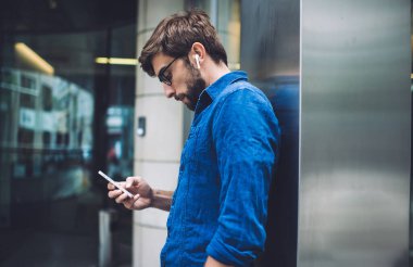 Gözlüklü, kulaklıklı ve kot gömlekli sakallı bir adamın sokakta metal sütuna yaslanırken cep telefonu kullanışının yan görüntüsü.