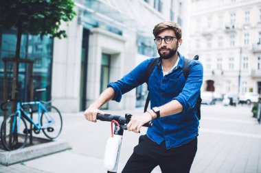 Gözlüklü, sırt çantalı ve sokağın ortasında elektrikli scooterla gezen neşeli ve sakallı bir erkek.