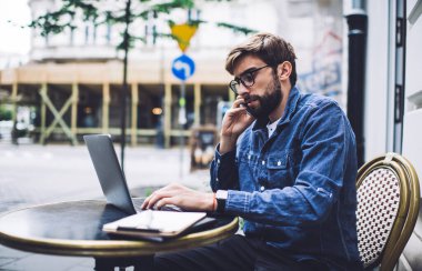 Kot ceketli, cep telefonuyla konuşan ve öğleden sonra siyah masada otururken netbook 'ta yazan uzaktan kumandalı erkek işçinin yan görüntüsü.