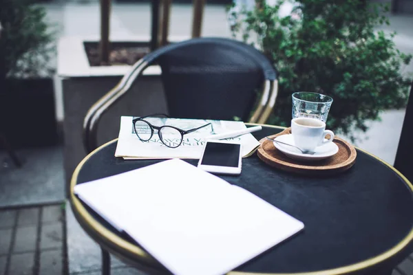 From above of cup of coffee with glass of water newspaper with eyeglasses and pen open notebook and smartphone on round black table in coffee shop