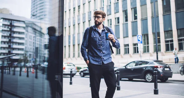 严肃的大胡子男嬉皮士身穿休闲装 戴着眼镜 在近现代建筑物的街道上散步 一边看着一边喝咖啡 — 图库照片