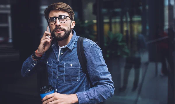 Pensive bearded male with coffee standing near glass door and talking on mobile phone while looking away and thinking near modern building